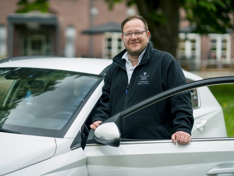 Ein Mitarbeiter des ambulanten Pflegedienstes lehnt an ihrem Dienstfahrzeug und lächtelt in die Kamera.