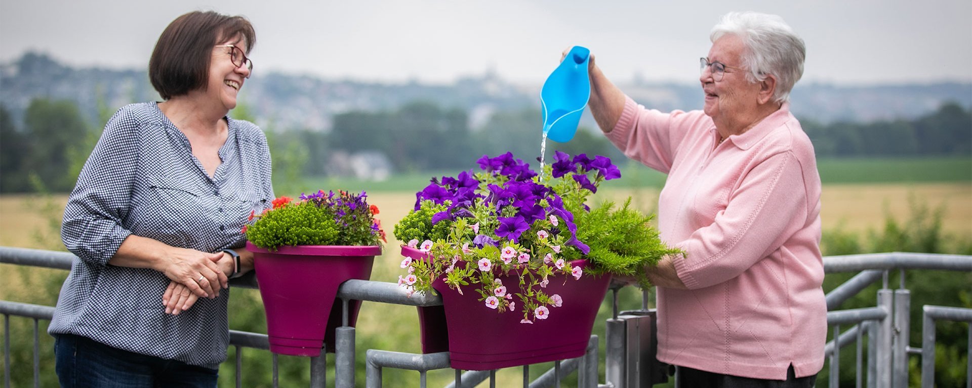 Zwei Frauen stehen an einem Geländer und gießen Blumen - lachen dabei